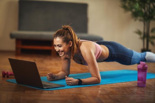 Young smiling woman is doing plank exercises in the living room on floor mat at home, looking at the laptop, in morning sunshine.