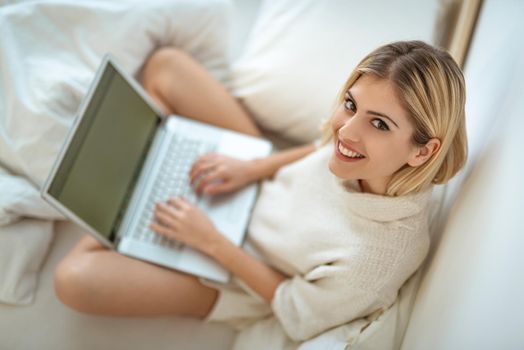 Beautiful young smiling woman relaxing in bed and surfing the net on laptop.