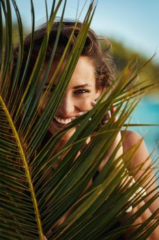 Portrait of a shy young woman is posing and looking under palm tree leaf at the tropical beach.
