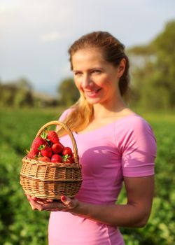 Basket full of red ripe strawberries, blurred young woman holding it, and strawberry fields in background.