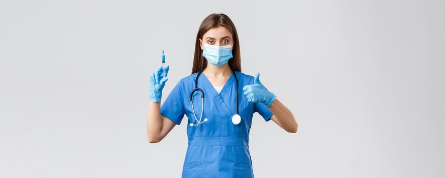 Covid-19, preventing virus, healthcare workers and quarantine concept. Confident female nurse, doctor in blue scrubs, medical mask and gloves assure in quality of coronavirus vaccine, hold syringe.