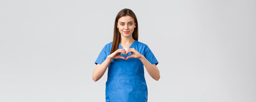 Healthcare workers, prevent virus, insurance and medicine concept. Smiling attractive female doctor, nurse in blue scrubs stay safe home, show heart sign, express respect to coronavirus fighters.