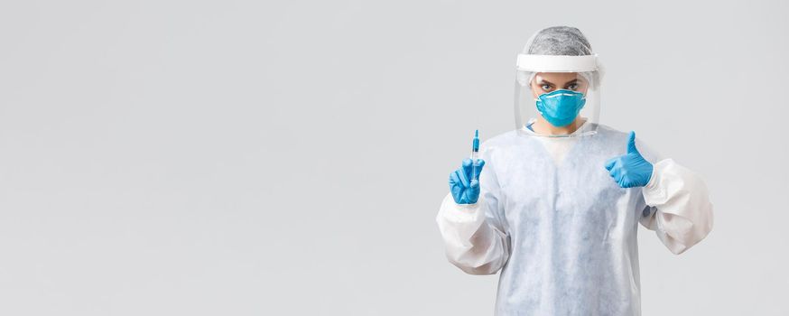 Covid-19, medical research, healthcare workers and quarantine concept. Confident professional doctor in personal protective equipment PPE costume, show thumb-up, hold syringe with corona vaccine.