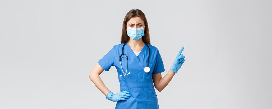Covid-19, preventing virus, health, healthcare workers and quarantine concept. Skeptical or doubtful female nurse in blue scrubs and medical mask, gloves pointing right, frowning disbelief.