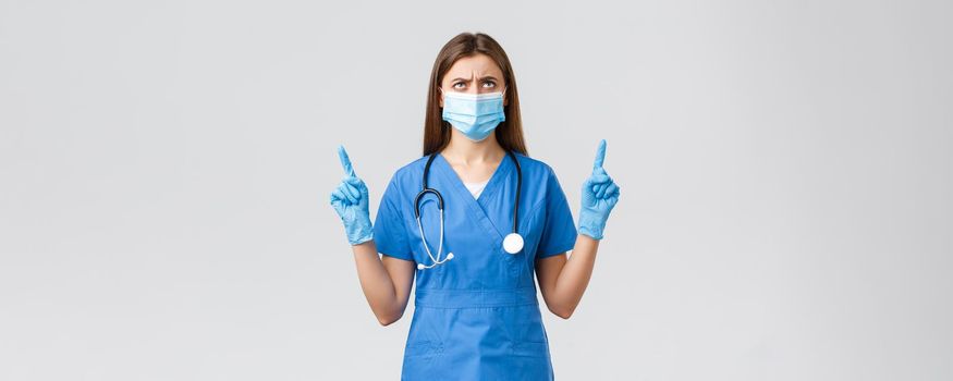 Covid-19, preventing virus, health, healthcare workers and quarantine concept. Concerned and disappointed female nurse or doctor in blue scrubs, medical mask, frowning looking up displeased.