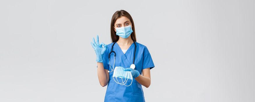 Covid-19, preventing virus, health, healthcare workers concept. Professional female nurse or doctor in blue scrubs and personal protective equipment, show okay sign and give medical mask.