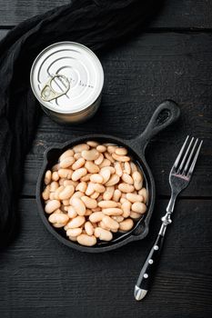 White kidney beans set, with metal can, in cast iron frying pan, on black wooden table background, top view flat lay