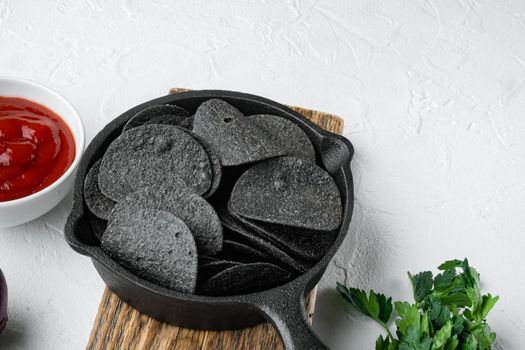 Black crispy potato chips set, with dipping sauces tomato dip sour cream, in cast iron frying pan, on white stone surface, with copy space for text