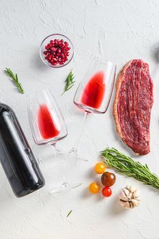 Two glasses of red wine near bottle and beef steaks over white concrete background, top view