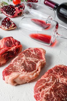 Raw set of alternative beef cuts Chuck eye roll, top blade, rump steak with red wine in glass and bottle, herbs and pomegranate. Organic meat. White textured background. Side view vertical