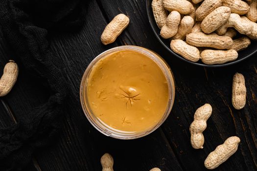 Peanut paste in an open jar and peanuts set, on black wooden table background, top view flat lay, with copy space for text