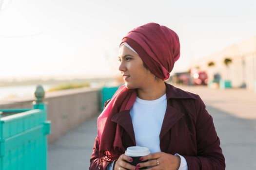 Young Arabic beautiful woman in hijab walking at street, tapping on smartphone and drinking coffee to-go. Pretty muslim female in headscarf strolling at aeroport, texting message and sipping drink.