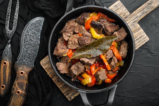 Beef meat stewed with potatoes, carrots and spices set, in cast iron frying pan, on black stone background, top view flat lay