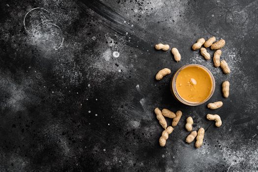 Peanut paste in an open jar and peanuts set, on black dark stone table background, top view flat lay, with copy space for text