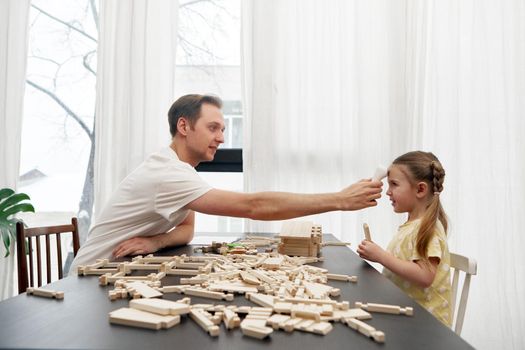 Side view of dad checking body temperature with infrared forehead thermometer of daughter sitting at table and playing with wooden blocks