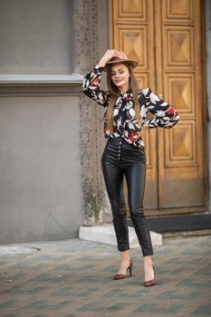 woman in shirt with flower print and leather pants posing to the camera near a grey building