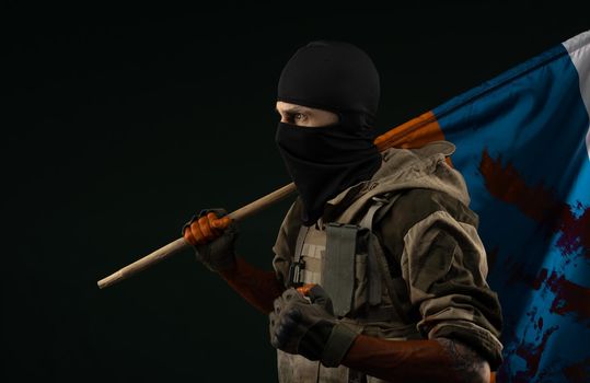 male soldier in a military uniform with a balaclava and hands covered in blood holds a bloody flag of Russia on a black background