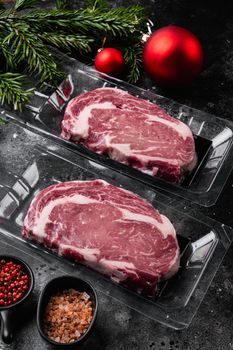 Rib-eye beef steak in plastic packing tray with Christmas tree decorations set, on black dark stone table background