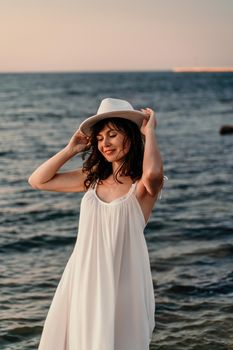 A woman in a white dress and hat is standing on the beach enjoying the sea. Happy summer holidays.