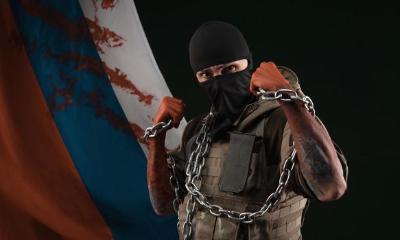 male soldier in a military uniform with a balaclava and hands covered in blood holds a chain as a symbol of slavery against the background of the bloody flag of Russia