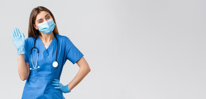 Covid-19, preventing virus, health, healthcare workers and quarantine concept. Friendly female nurse in clinic wearing blue scrubs and medical mask, waving hello or hi sign with gloves on.