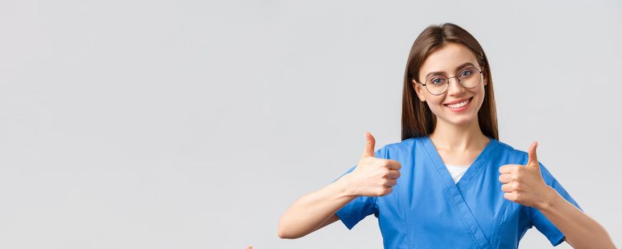 Healthcare workers, medicine, insurance and covid-19 pandemic concept. Excited happy female nurse in blue scrubs, doctor show thumbs-up in support or approval, encourage make tests in clinic.