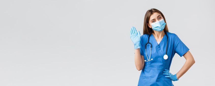 Covid-19, preventing virus, health, healthcare workers and quarantine concept. Friendly female nurse in clinic wearing blue scrubs and medical mask, waving hello or hi sign with gloves on.