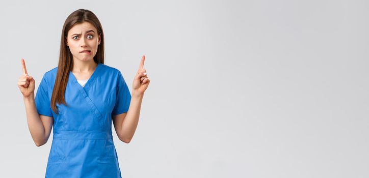 Healthcare workers, prevent virus, insurance and medicine concept. Worried or nervous nurse or doctor in blue scrubs, biting lip concerned, looking up, cant resist temptation, grey background.