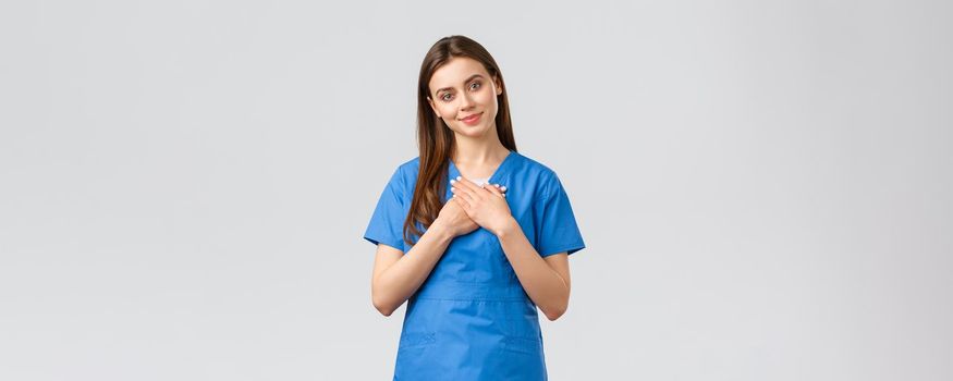 Healthcare workers, prevent virus, insurance and medicine concept. Touched and flattered female nurse or doctor in blue scrubs, tilt head and touch heart, smile from compliment, nice gesture.