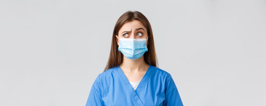 Covid-19, preventing virus, healthcare workers and quarantine concept. Skeptical female nurse or doctor in blue scrubs, medical mask, raise one eyebrow intrigued and confused, look upper left corner.