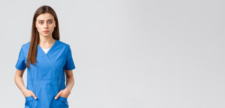 Healthcare workers, prevent virus, insurance and medicine concept. Serious-looking, confident young medical worker, nurse or doctor in blue scrubs hold hands in pockets and looking camera.