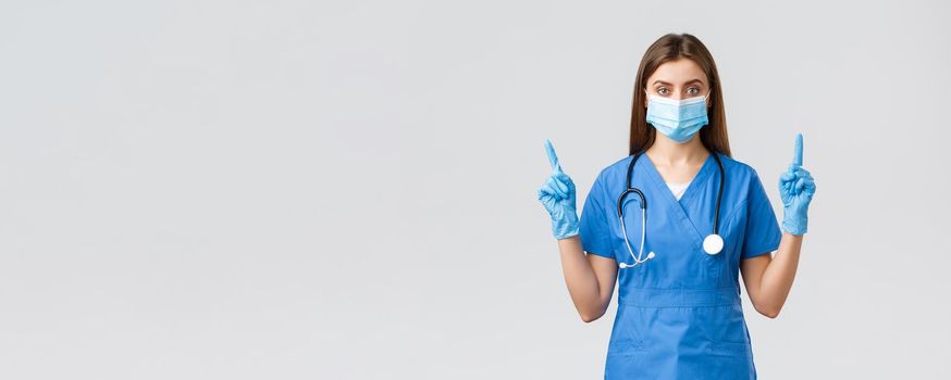 Covid-19, preventing virus, health, healthcare workers concept. Serious and confident female nurse in blue scrubs, medical mask PPE, pointing fingers up, inform patients how prevent corona infection.