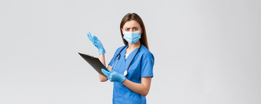 Covid-19, preventing virus, health, healthcare workers and quarantine concept. Displeased and bothered female nurse or doctor in blue scrubs and medical mask, reading nonsense on clipboard.