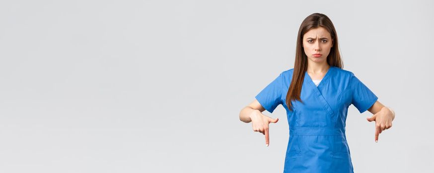 Healthcare workers, prevent virus, covid-19 test screening, medicine concept. Disappointed and uneasy cute nurse or doctor in blue scrubs, pointing fingers down, showing bad unpleasant news.