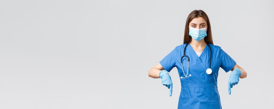 Covid-19, preventing virus, health, healthcare workers and quarantine concept. Determined female nurse or doctor in blue scrubs, medical mask and gloves, pointing fingers down, showing info banner.