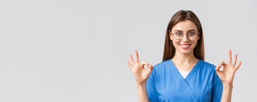 Healthcare workers, medicine, insurance and covid-19 pandemic concept. Confident and optimistic female nurse, doctor in blue scrubs and glasses, show okay sign, guarantee best service quality.