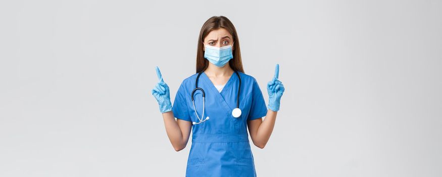 Covid-19, preventing virus, health, healthcare workers and quarantine concept. Skeptical female nurse in blue scrubs and medical mask dont believe this info, pointing fingers up doubtful.