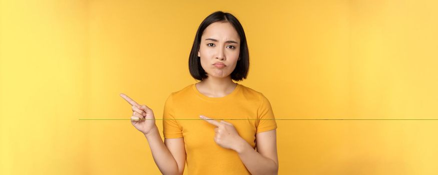 Upset asian woman pointing fingers left, showing smth disappointing, complaining at banner, standing over yellow background.