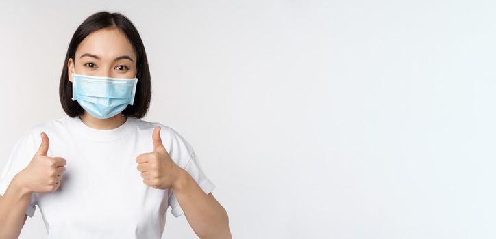 Covid-19, health and people concept. Young happy korean woman in medical mask, showing thumbs up, standing in tshirt over white background.