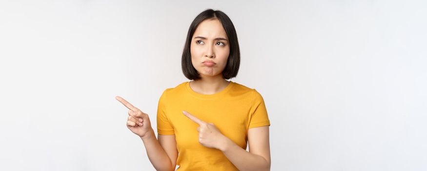 Upset grumpy asian girl, pouting and looking at smth unfair, pointing fingers left at banner, logo brand, standing in yellow tshirt over white background.