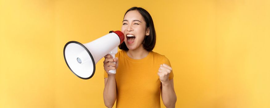 Announcement. Happy asian woman shouting loud at megaphone, recruiting, protesting with speaker in hands, standing over yellow background.