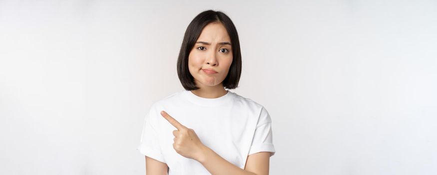 Skeptical asian girl in white t-shirt, pointing at product or logo with disappointed grimace, dislike and complain at smth, standing over white background.