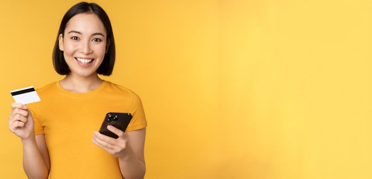 Online shopping. Cheerful asian girl holding credit card and smartphone, paying, order with mobile phone, standing over yellow background.