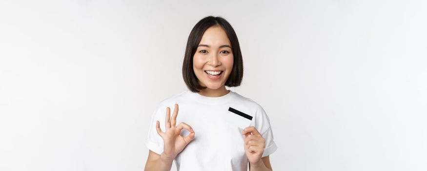 Smiling korean girl showing okay sign and recommending credit card of copy space bank, standing in tshirt over white background.