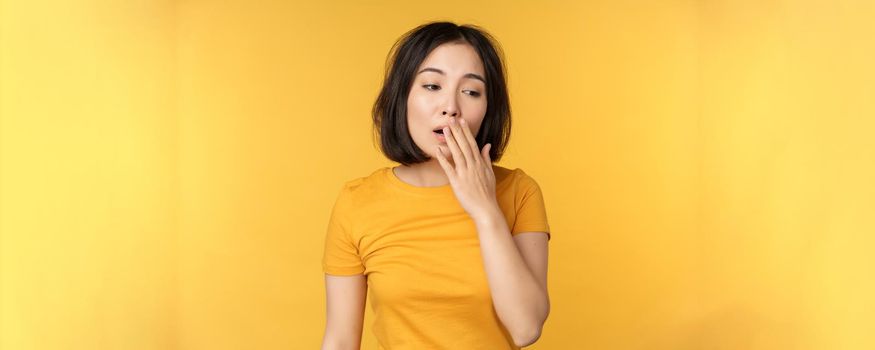 Image of young asian girl student, yawning, feeling tired, standing sleepy agaisnt yellow background. Copy space