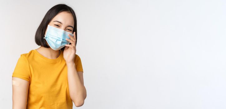 Covid-19, vaccination and healthcare concept. Cute asian girl in medical face mask, showing band aid after coronavirus vaccination, standing over white background.