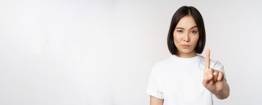 Image of asian girl showing stop, prohibit smth, extend one arm to show forbidding, taboo gesture, standing in tshirt over white background.