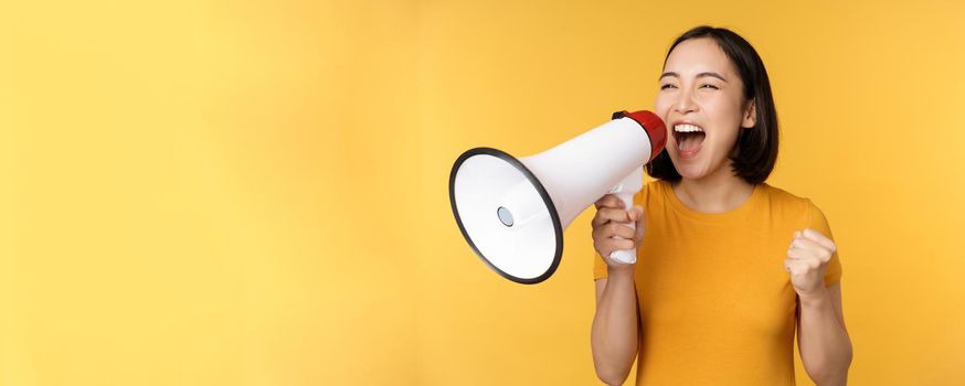 Announcement. Happy asian woman shouting loud at megaphone, recruiting, protesting with speaker in hands, standing over yellow background.