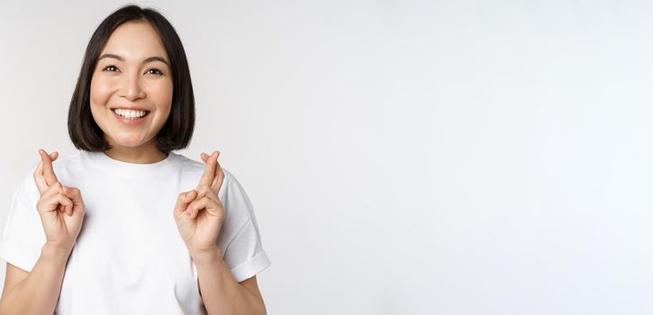 Hopeful asian girl cross fingers, making wish, anticipating, wishing smth, standing in tshirt over white background. Copy space