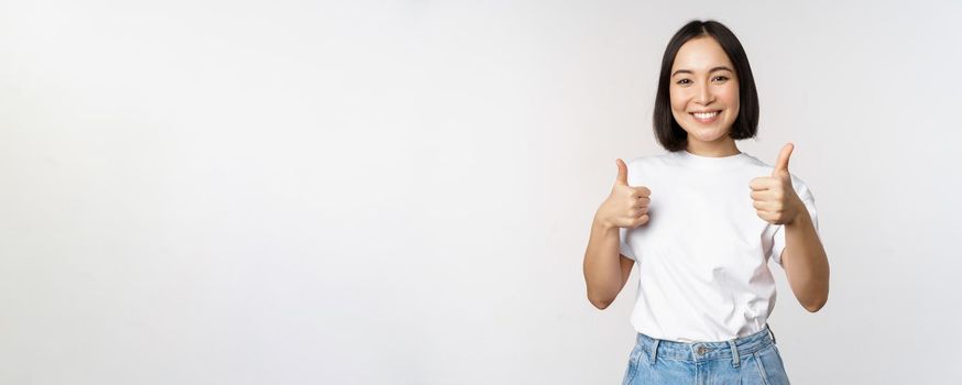 Happy beautiful korean girl, showing thumbs up in approval, smiling pleased, like smth, recommending, standing over white background.